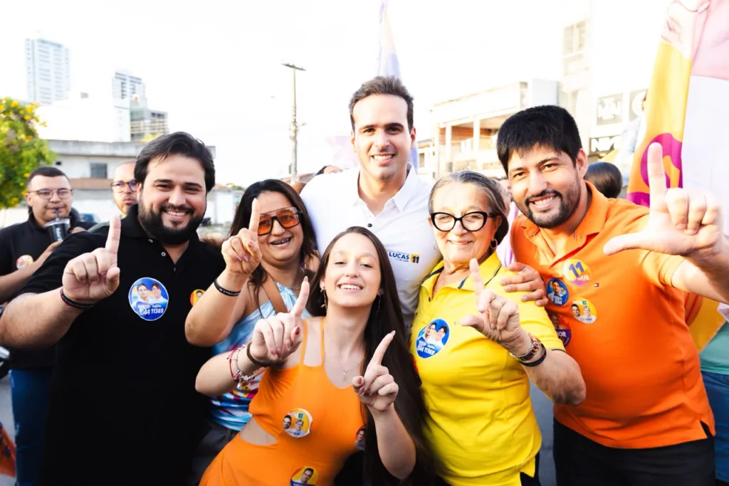 Lucas Ribeiro sorri em plano médio cercado por apoiadores fazendo o número um com os dedos durante o dia.