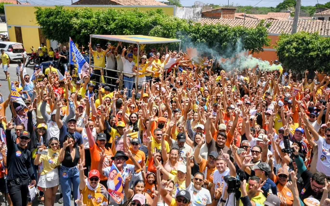 Multidão reunida na rua com braços levantados e sinal de campanha em frente a veículo com comitiva e fumaça colorida.