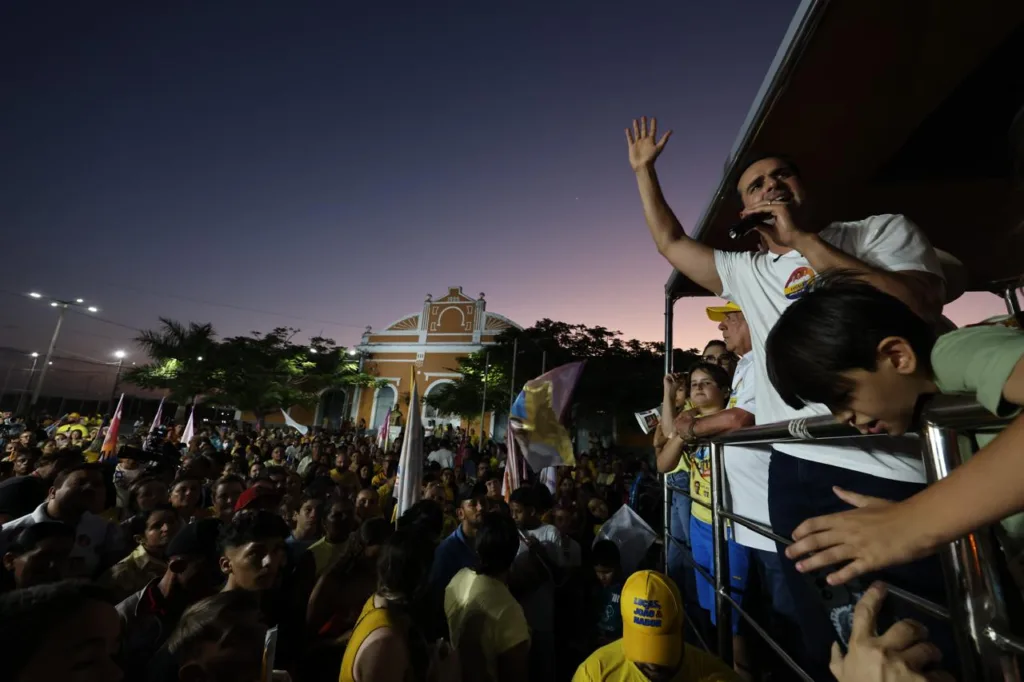 Lucas Ribeiro discursa ao microfone com o braço erguido no veículo de campanha ao entardecer, diante de grande público.