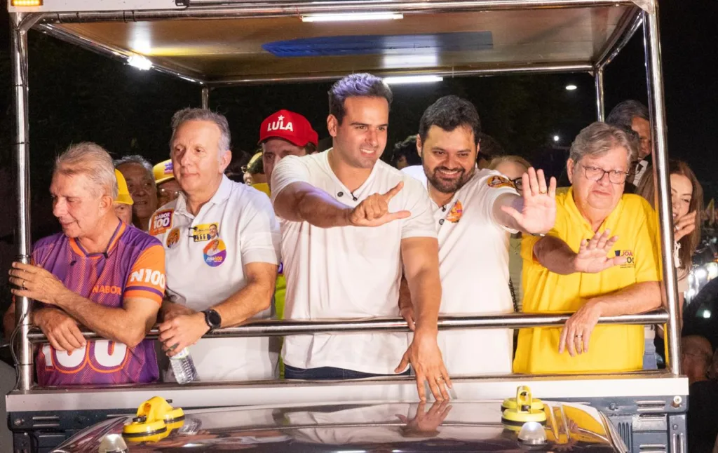 Lucas Ribeiro faz gesto de campanha ao lado de Aguinaldo Ribeiro, João Azevêdo e lideranças em veículo aberto.