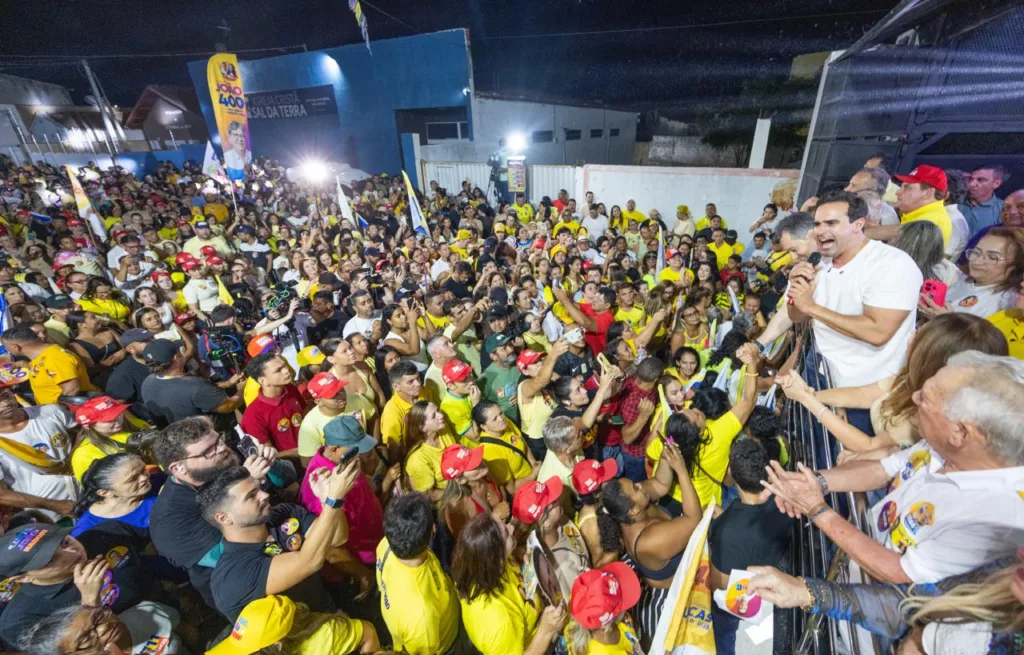 Vista superior de Lucas Ribeiro discursando ao microfone inclinado sobre a barra do veículo para a multidão abaixo.