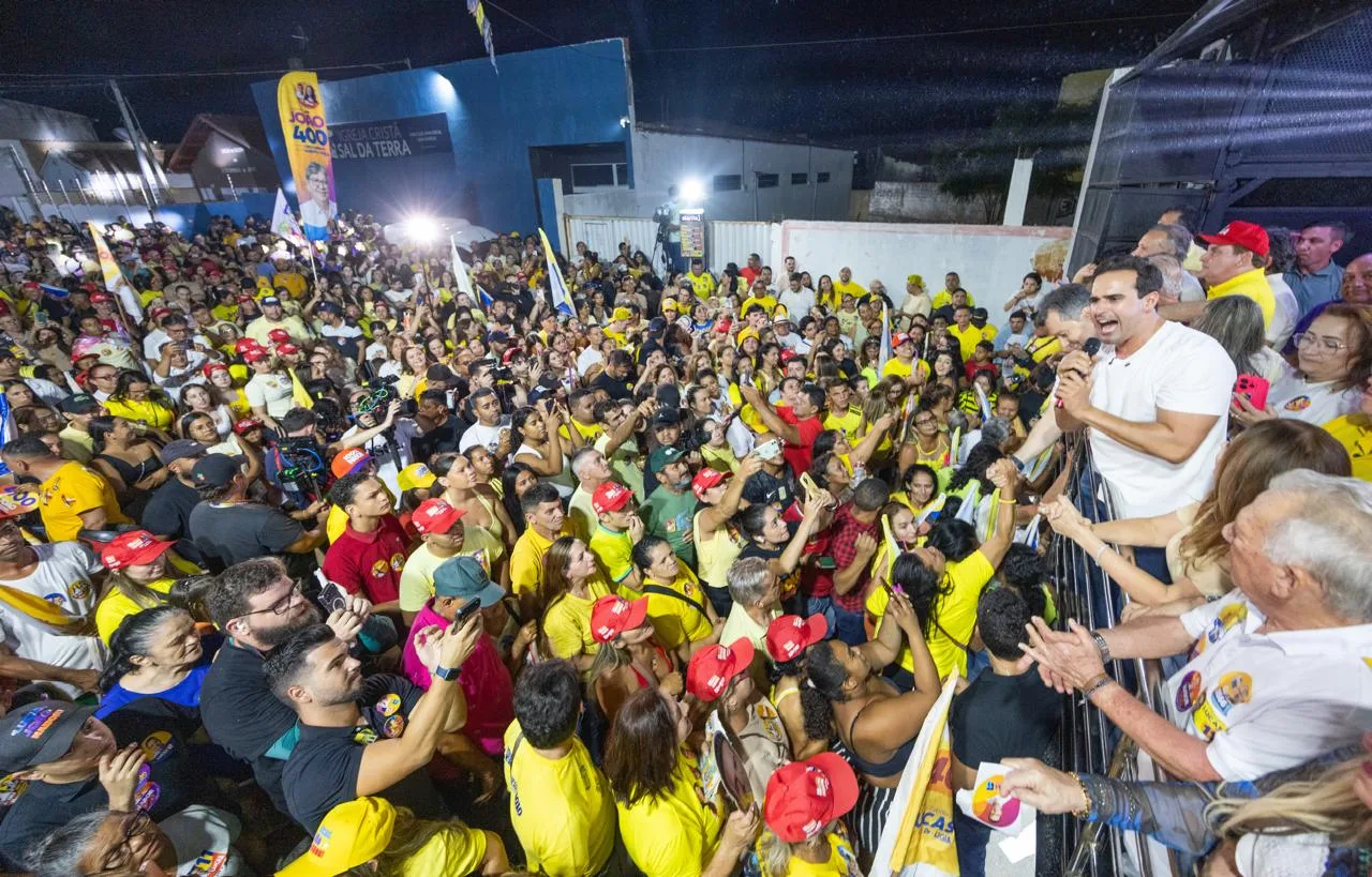 Vista superior de Lucas Ribeiro discursando ao microfone inclinado sobre a barra do veículo para a multidão abaixo.