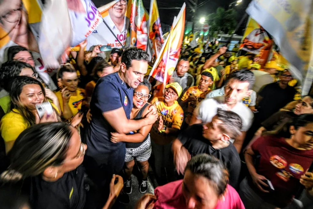 Lucas Ribeiro de camisa escura abraça moradora sorridente em meio à militância com bandeiras ao redor.