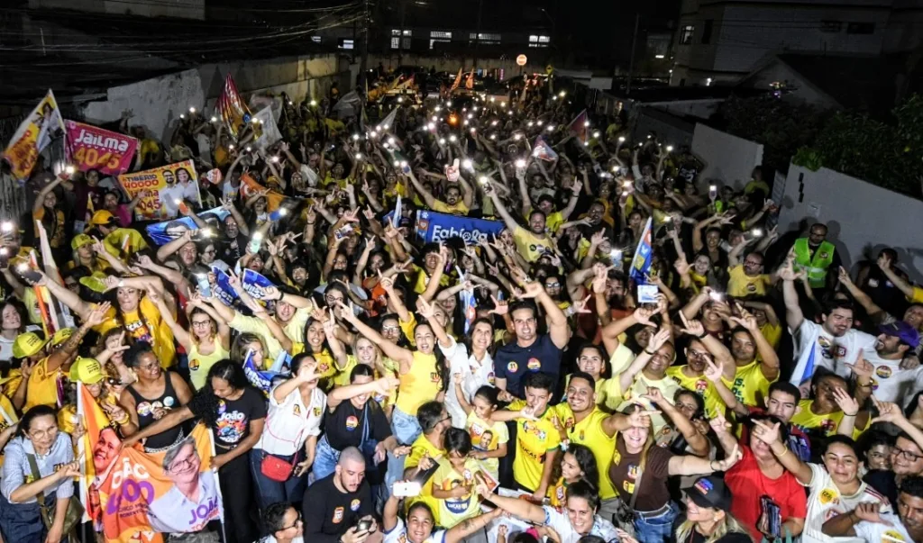 Imagem aérea noturna de multidão com celulares iluminados e bandeiras acompanhando Lucas Ribeiro e comitiva em rua residencial.