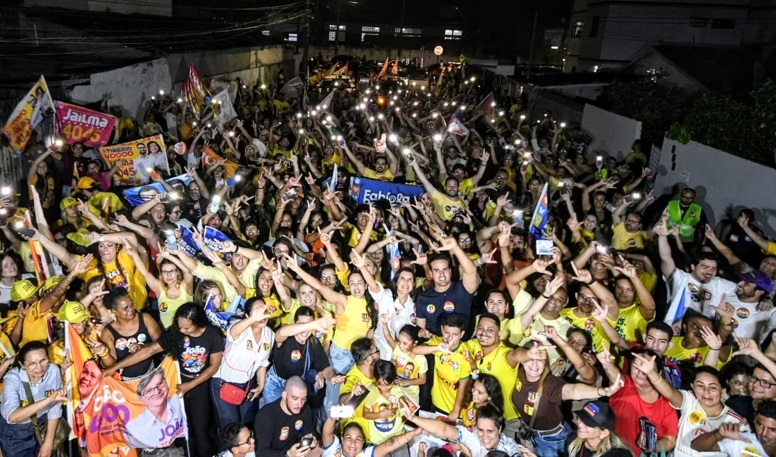 Imagem aérea noturna de multidão com celulares iluminados e bandeiras acompanhando Lucas Ribeiro e comitiva em rua residencial.