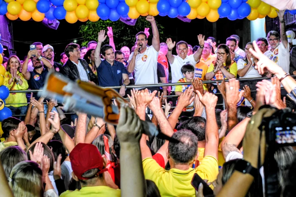 Lucas Ribeiro ao microfone com braço erguido no centro do palco cercado por Adriano Galdino, Murilo Galdino e lideranças sob balões amarelos e azuis.