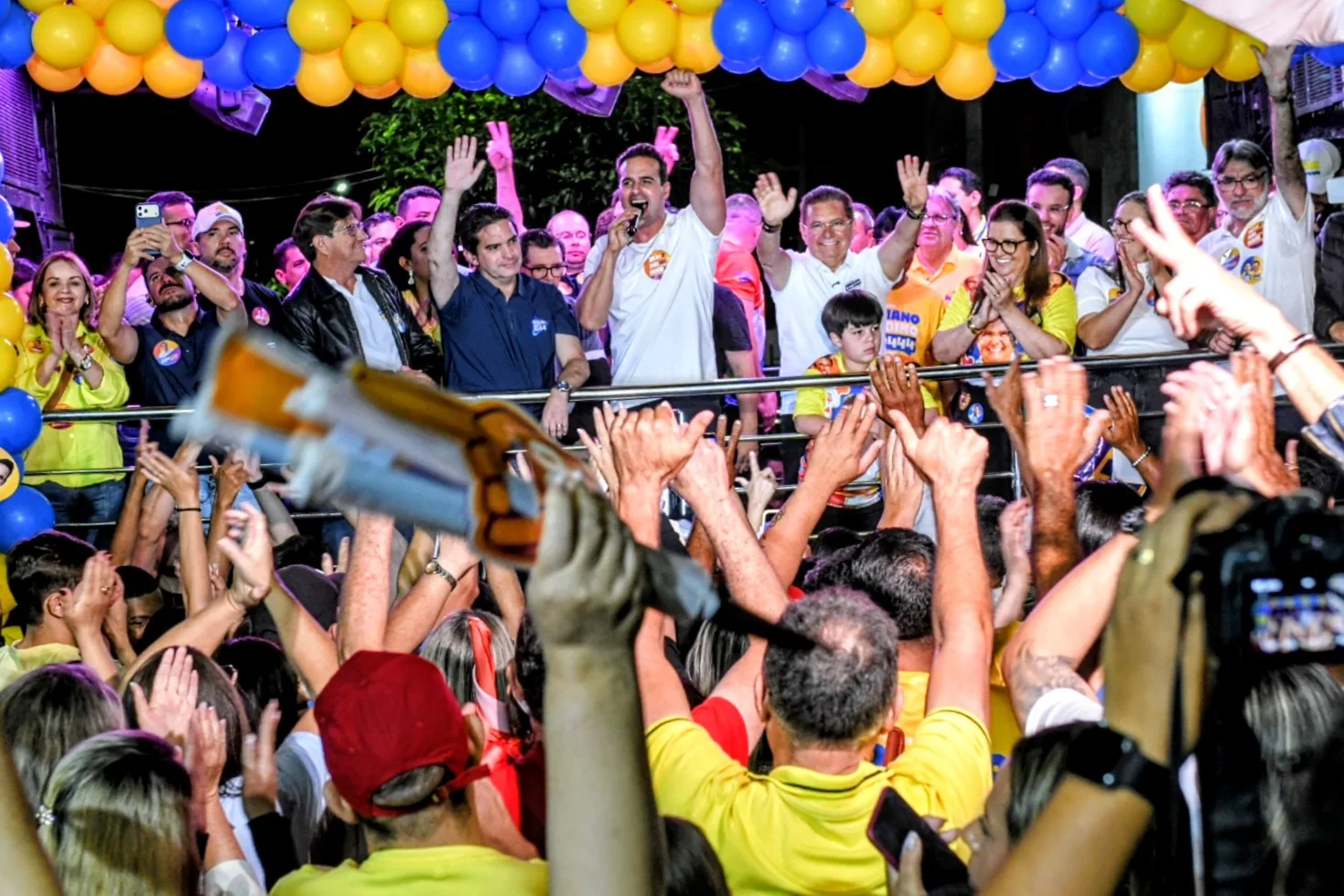 Lucas Ribeiro ao microfone com braço erguido no centro do palco cercado por Adriano Galdino, Murilo Galdino e lideranças sob balões amarelos e azuis.