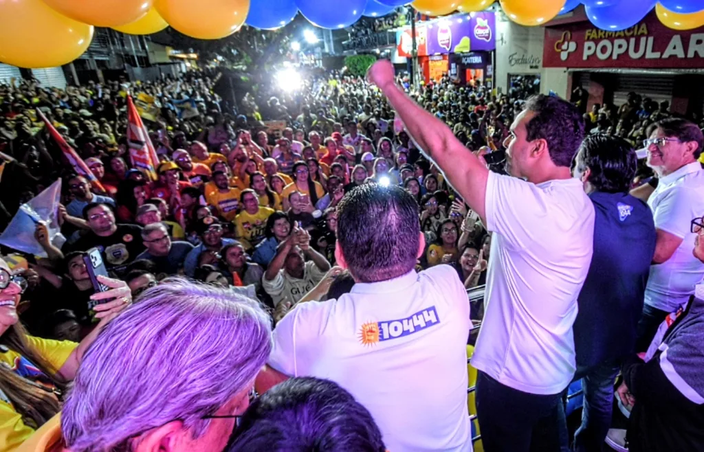 Lucas Ribeiro veste camisa branca e ergue o punho ao microfone do alto do palanque diante de grande público na rua.