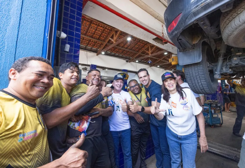 Lucas Ribeiro, Lígia Feliciano e Nabor Wanderley posam sorridentes ao lado de mecânicos que fazem sinal de positivo sob carro no elevador.