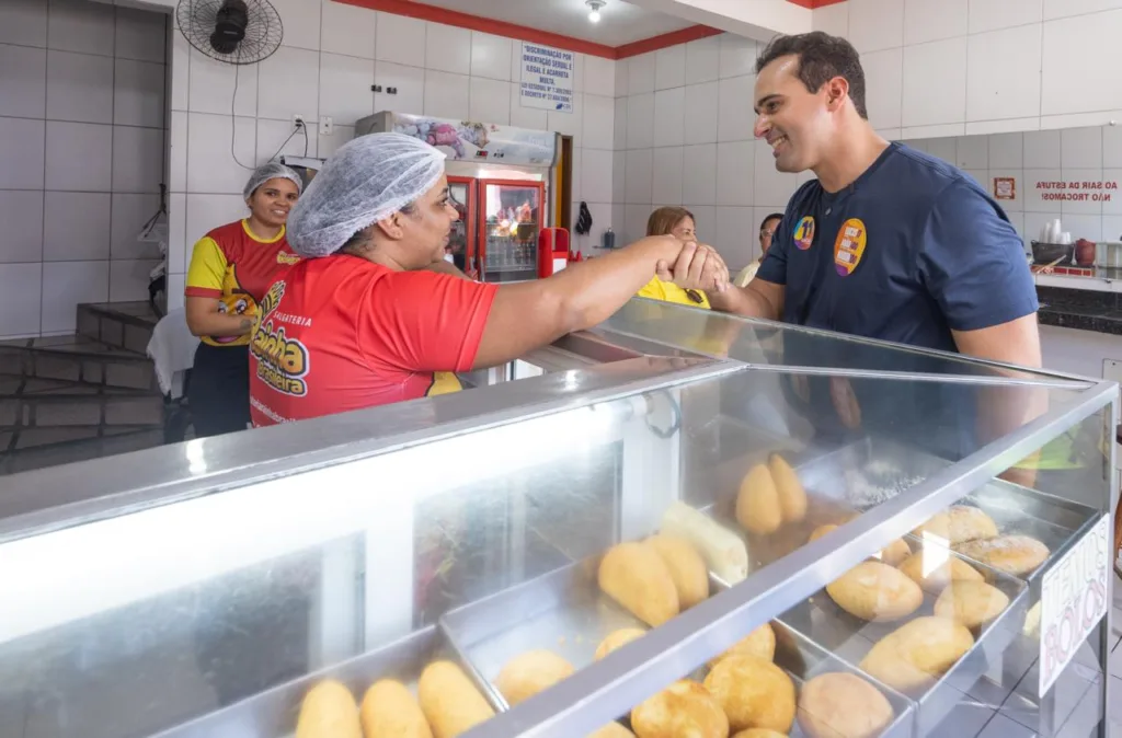 Lucas Ribeiro sorri e cumprimenta funcionária de touca atrás do balcão de vidro com salgados expostos.